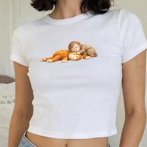 May include: White crop top featuring a graphic of two cartoon monkeys. One monkey is brown and orange, and the other is brown and sleeping on top of the first. The shirt has short sleeves and a crew neck.