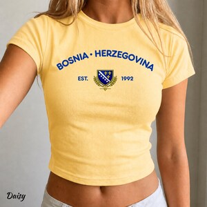 Pu&ograve; includere: Un top corto giallo con la scritta blu "BOSNIA &bull; HERZEGOVINA" ad arco sopra uno stemma. Lo stemma presenta un disegno blu e bianco con dettagli dorati e la scritta "EST. 1992".