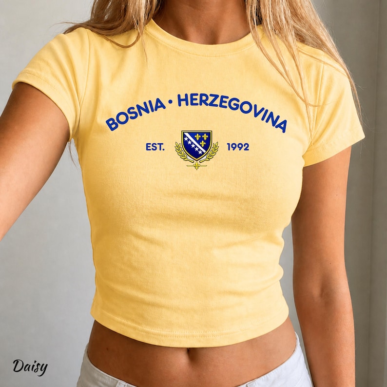Pu&ograve; includere: Un top corto giallo con la scritta blu "BOSNIA &bull; HERZEGOVINA" ad arco sopra uno stemma. Lo stemma presenta un disegno blu e bianco con dettagli dorati e la scritta "EST. 1992".