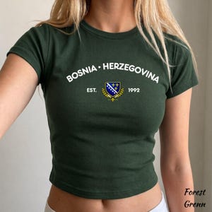 Pu&ograve; includere: T-shirt verde scuro Forest Grenn con la scritta bianca "BOSNIA &bull; HERZEGOVINA". Sotto, uno stemma con la bandiera bosniaca e la scritta "EST. 1992". La maglietta &egrave; un crop top a maniche corte.