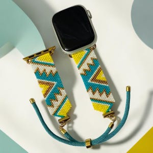 May include: A beaded Apple Watch band with a geometric pattern in yellow, turquoise, white, and gold. The band is connected to a silver Apple Watch. A teal cord with gold accents is attached to the band.