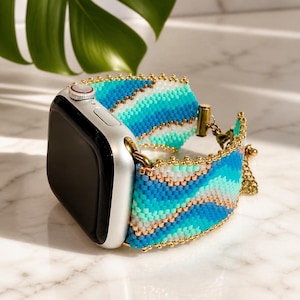 May include: A silver smartwatch featuring a beaded watch band with a wavy pattern in shades of turquoise, blue, and white, accented with metallic gold beads. The band includes a gold-toned metal chain and clasp mechanism for secure attachment.