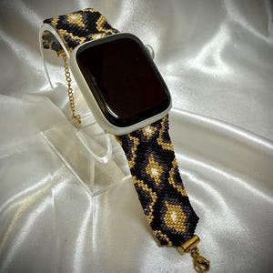 May include: A smartwatch with a rectangular black screen and a silver casing, featuring a custom beaded watch band. The band displays a repeating diamond pattern in black, gold, and white beads, finished with gold-toned metal hardware and a clasp.