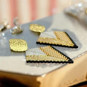 May include: Close-up of gold, white, and black beaded earrings. The earrings have a geometric design with a triangular shape. Two pairs of earrings are visible, along with two small gold textured studs.
