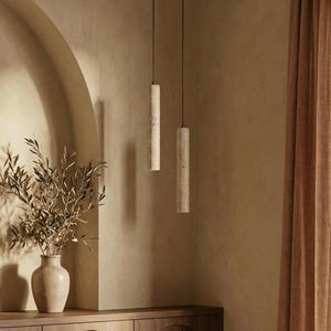 May include: Two cylindrical pendant lights hang from the ceiling in a room with a warm, neutral colour palette. The lights are made of a textured, light-coloured material. A vase with dried branches sits on a wooden cabinet.