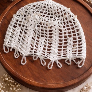 May include: A white, crocheted head covering with a delicate open weave design. The head covering is adorned with small, round pearls and features a scalloped edge with teardrop-shaped pearl accents. It rests on a wooden tray.
