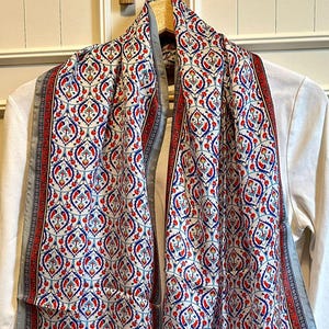 May include: A patterned scarf with a white background and a repeating design in red and blue. The scarf has a gray border and is draped over a white long-sleeved shirt. The scarf is made of a lightweight fabric.