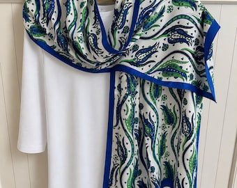 May include: A white long-sleeve shirt draped with a rectangular scarf featuring a blue, green, and white tulip pattern. The scarf has a solid blue border and a small white label in the corner with text that reads "Silk & Cashmere Made in Turkey".