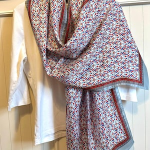 May include: A patterned scarf with a white background and a repeating design in red, blue, and gray. The scarf is draped over a white long-sleeved shirt. The scarf has a light blue border.