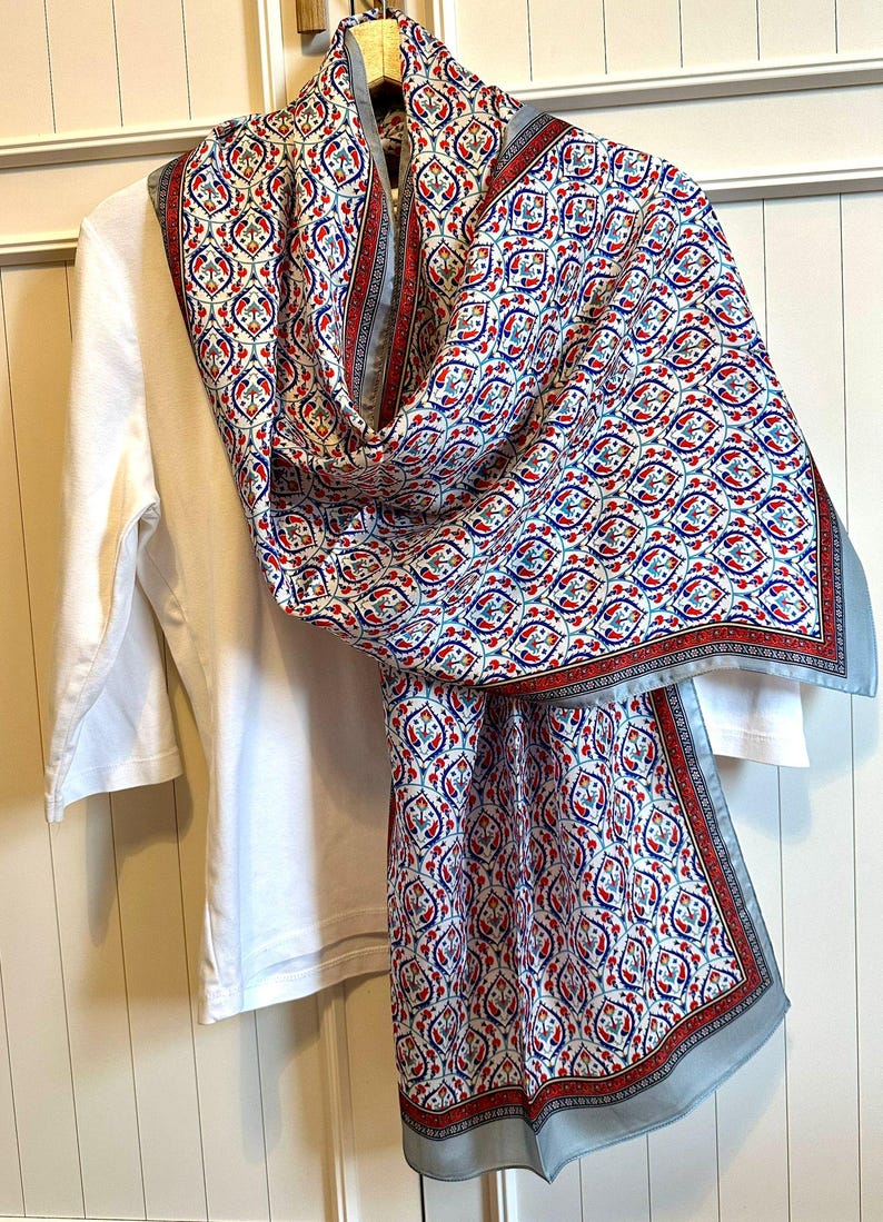 May include: A patterned scarf with a white background and a repeating design in red, blue, and gray. The scarf is draped over a white long-sleeved shirt. The scarf has a light blue border.