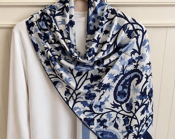 May include: A white long-sleeve shirt draped with a large square scarf featuring a blue and white floral and paisley pattern. The scarf has a dark blue border with a striped section at the bottom corner, contrasting against the plain white shirt.
