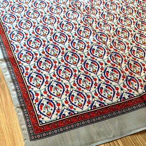 May include: A decorative scarf with a repeating pattern of red, blue, and teal floral motifs on a cream background. The scarf has a red border with black and white accents and a light gray border.