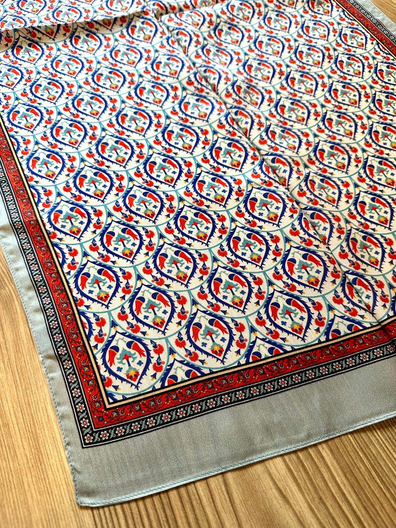 May include: A decorative scarf with a repeating pattern of red, blue, and teal floral motifs on a cream background. The scarf has a red border with black and white accents and a light gray border.