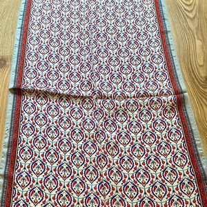 May include: A long, rectangular scarf with a repeating pattern of red, blue, and white floral motifs on an off-white background. The scarf has a light blue border with a red and white patterned trim.