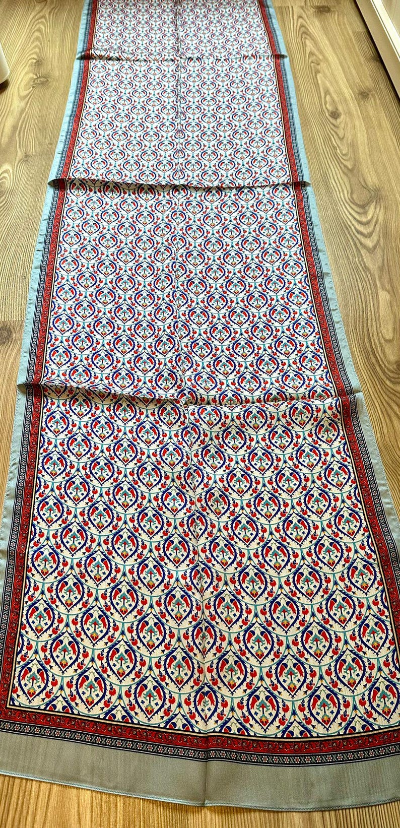 May include: A long, rectangular scarf with a repeating pattern of red, blue, and white floral motifs on an off-white background. The scarf has a light blue border with a red and white patterned trim.