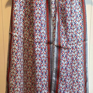 May include: A patterned scarf with a white background and a repeating design of red and blue floral motifs. The scarf has a red border with a black and white pattern and a light gray trim. The scarf is hanging on a wooden hanger.