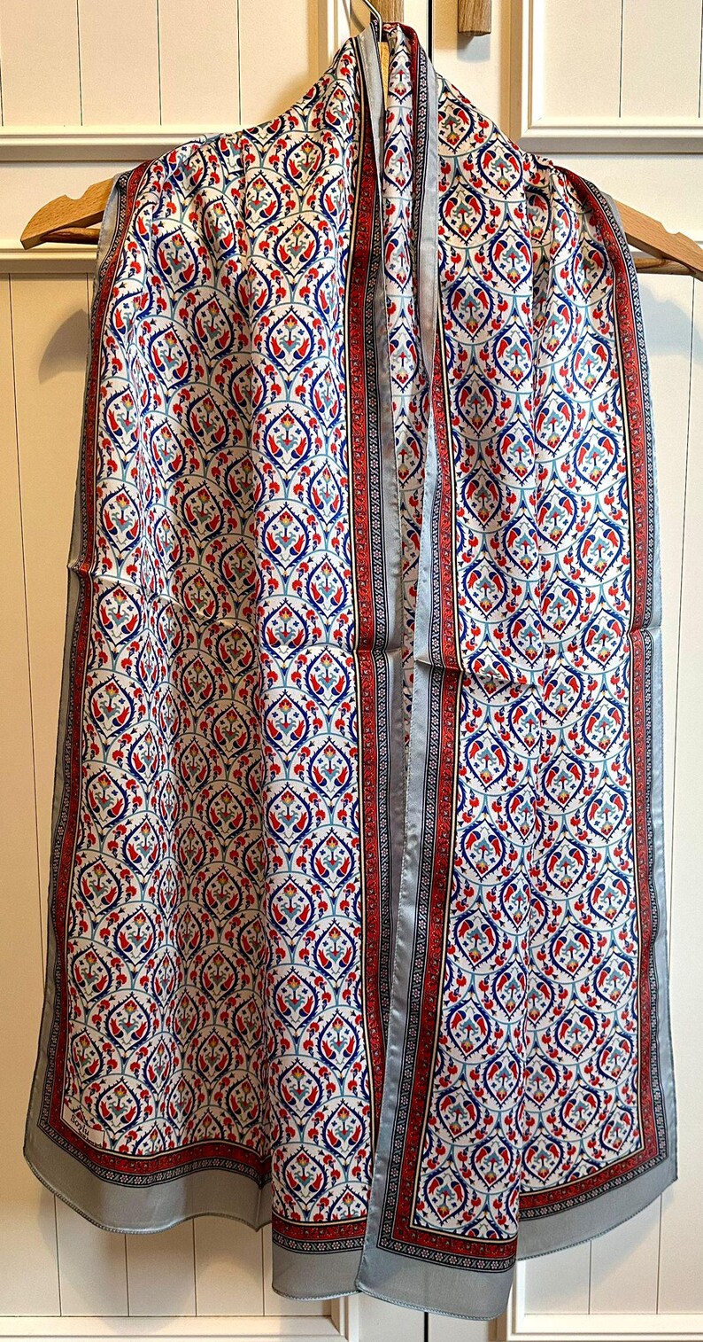 May include: A patterned scarf with a white background and a repeating design of red and blue floral motifs. The scarf has a red border with a black and white pattern and a light gray trim. The scarf is hanging on a wooden hanger.
