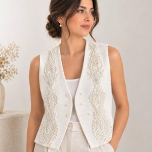 May include: A sleeveless white waistcoat featuring intricate vertical panels of white beadwork and embroidery along the front opening. The garment has a V-neckline and is adorned with small, round white buttons down the centre front, layered over a plain white top.