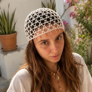 May include: Cream-colored crocheted head covering with a geometric pattern. The head covering has a beaded fringe in shades of pink, orange, and yellow. The head covering is worn by a person with long brown hair.