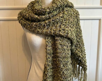 May include: A green and beige woven scarf with fringe. The scarf is draped over a mannequin, showcasing its texture and design. The scarf's color palette includes shades of green and beige, with a textured weave and fringe detail.