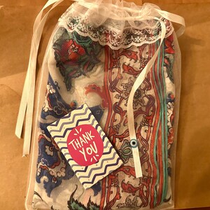 May include: A sheer mesh bag with a white lace trim, containing a patterned textile. A small card with the text "Thank You" is visible. The textile features a mix of red, blue, and white floral and paisley designs. The bag is tied with white ribbons.
