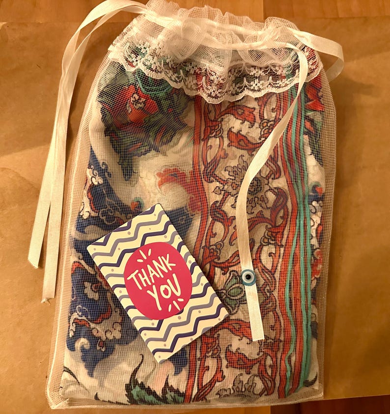 May include: A sheer mesh bag with a white lace trim, containing a patterned textile. A small card with the text "Thank You" is visible. The textile features a mix of red, blue, and white floral and paisley designs. The bag is tied with white ribbons.