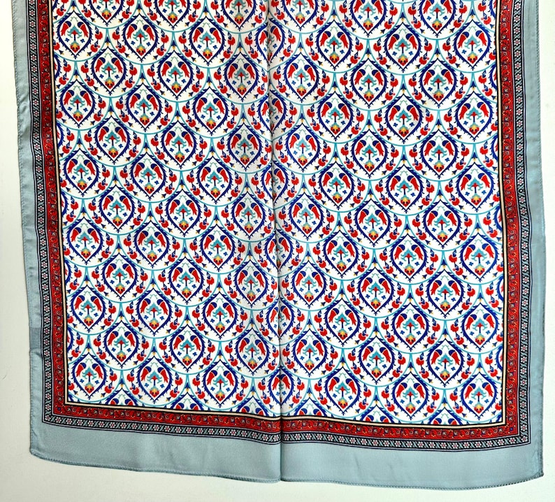 May include: A square scarf with a repeating pattern of blue, red, and turquoise floral motifs on a white background. The border features a red and black design, and the scarf has a light blue border.