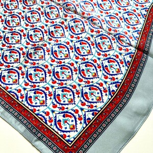 May include: A square silk scarf with a detailed pattern. The design features a repeating motif of stylized birds and floral elements in red, blue, and white. The border is a contrasting red and black floral pattern, with a light blue edge.