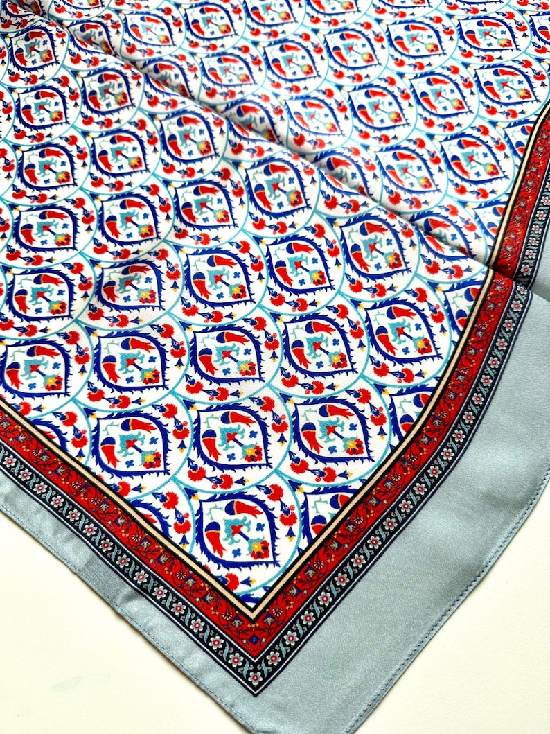 May include: A square silk scarf with a detailed pattern. The design features a repeating motif of stylized birds and floral elements in red, blue, and white. The border is a contrasting red and black floral pattern, with a light blue edge.