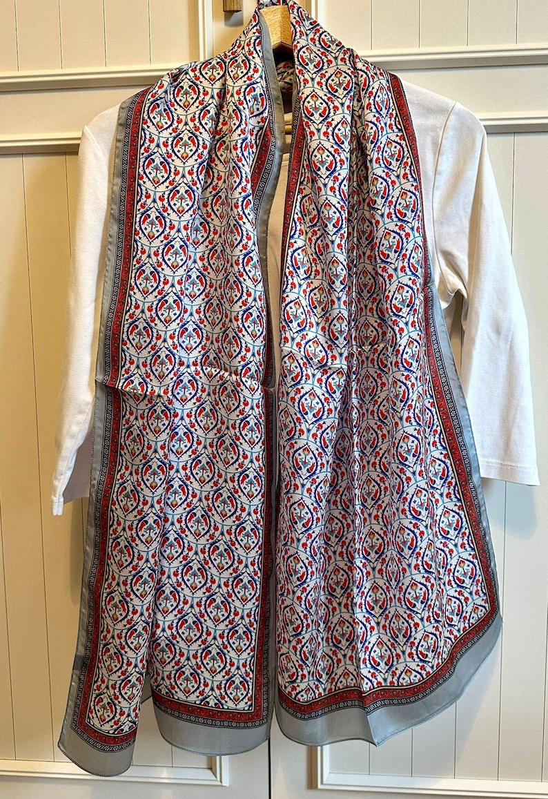 May include: A patterned scarf with a white background and a repeating design in red and blue. The scarf has a gray border and is draped over a white long-sleeved shirt. The scarf is made of a lightweight fabric.