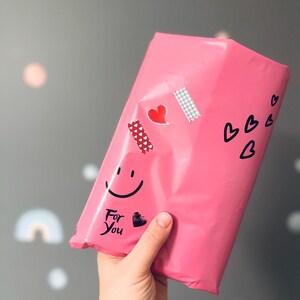 May include: A bright pink package decorated with heart stickers, a smiley face, and the words "For You." The package is held by a hand against a gray background with soft, colorful shapes.