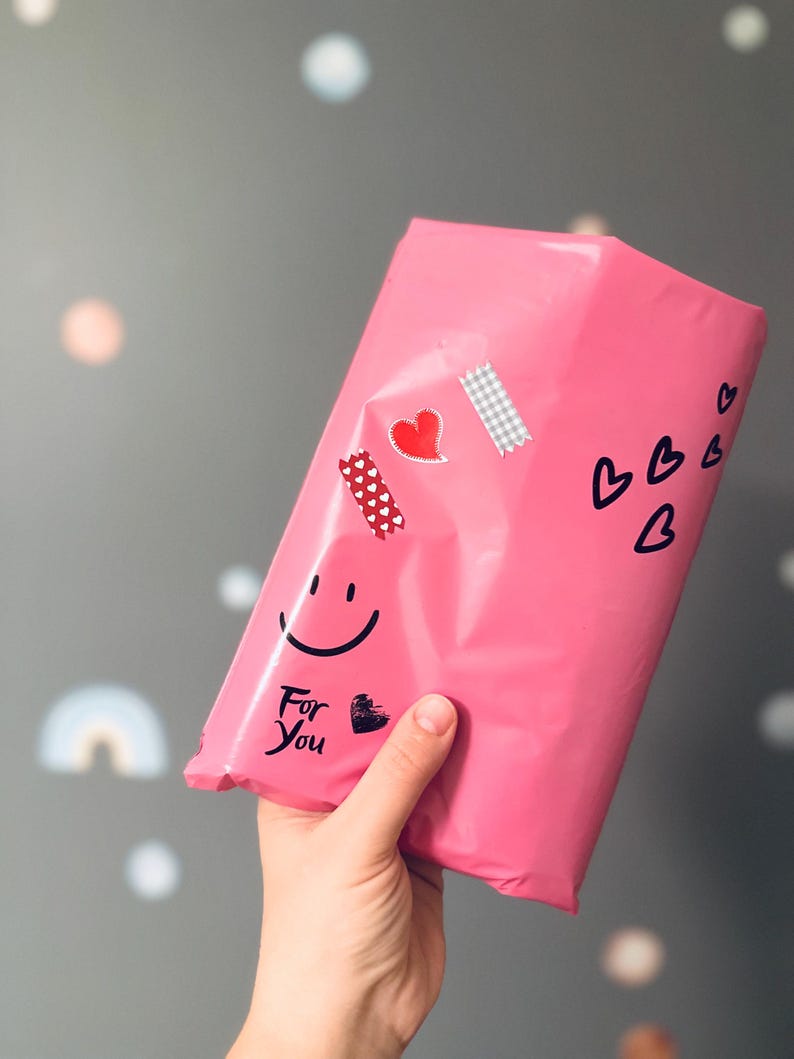 May include: A bright pink package decorated with heart stickers, a smiley face, and the words "For You." The package is held by a hand against a gray background with soft, colorful shapes.