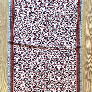May include: A rectangular scarf with a repeating pattern of red, blue, and white floral motifs. The scarf has a gray border and is displayed on a wooden surface. The scarf is approximately 60 inches long.