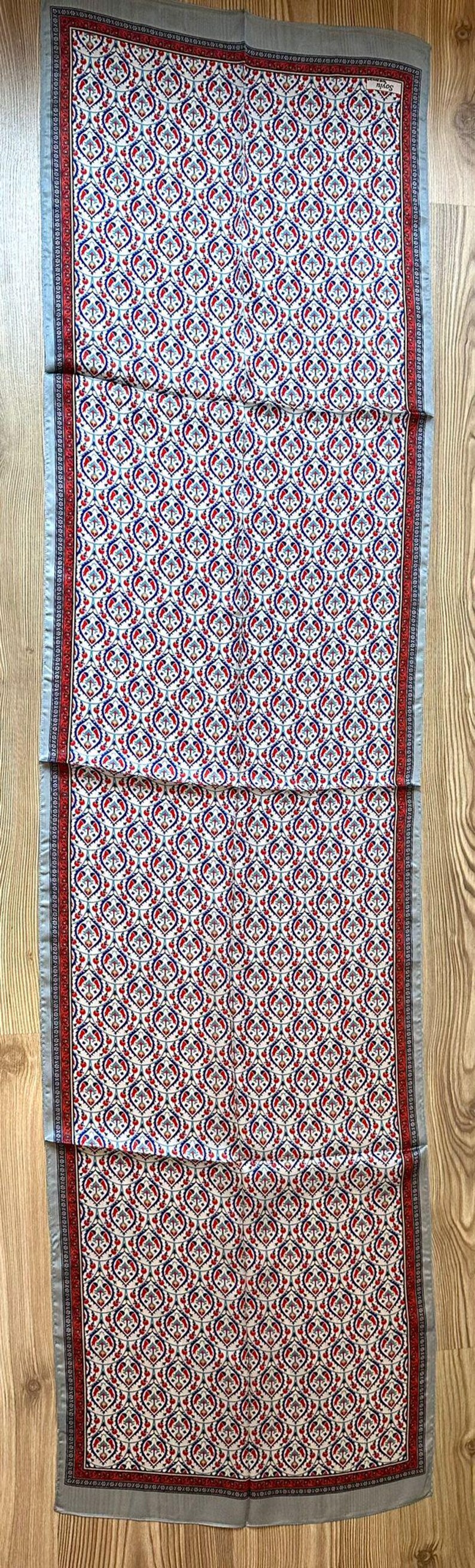 May include: A rectangular scarf with a repeating pattern of red, blue, and white floral motifs. The scarf has a gray border and is displayed on a wooden surface. The scarf is approximately 60 inches long.