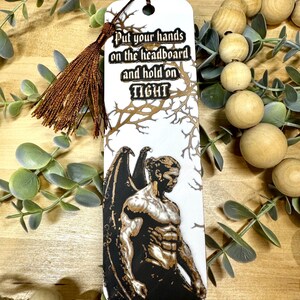 May include: White bookmark with a brown tassel, featuring a muscular figure with wings. The text "Put your hands on the headboard and hold on TIGHT" is printed on the bookmark. The design includes a tree branch pattern.