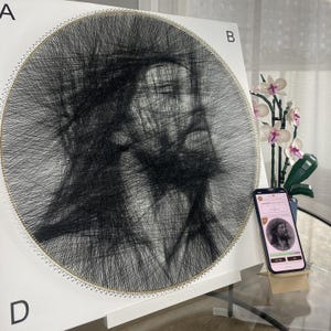 Może przedstawiać: Okrągły portret wykonany techniką string art z czarnej nici owiniętej wokół białej tablicy ze złotymi szpilkami. Litery A, B i D są nadrukowane w rogach. Smartfon wyświetla cyfrowy obraz przedstawiający mężczyznę z brodą.
