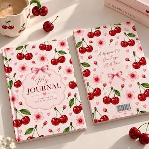 May include: Two journals with a light pink cover featuring a pattern of red cherries, pink cherry blossoms, and small hearts. The left journal displays the text My Journal A Happier Me One Page At A Time. The right journal displays A Happier Me One Page At A Time.