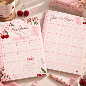 May include: Two pink and white planners with cherry and floral illustrations. The left planner is titled My Goals and the right planner is titled Year at a Glance. Both feature checkboxes, heart icons, and motivational text like You Can Do This.