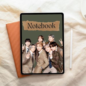 May include: A notebook with a green cover featuring a group of seven people in suits. The word "Notebook" is printed on a brown paper strip. An orange notebook and a white stylus are next to the notebook.