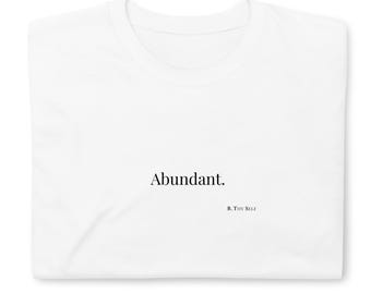 May include: A folded white t-shirt with the word "Abundant" printed in black, centered on the front. Below the word is the text "B. THY SELF" in a smaller font.