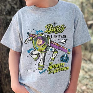 May include: A light gray t-shirt featuring a colorful graphic of Buzz Lightyear in his spacesuit. The design includes the words "Buzz Lightyear" and "To Infinity and Beyond" in a playful font, with a planet and stars.