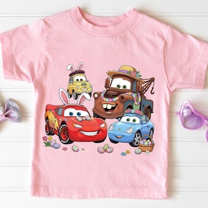 Può includere: Una maglietta rosa chiaro con una grafica di auto animate, tra cui un'auto da corsa rossa, un carro attrezzi marrone, un'auto blu e un'auto gialla. Le auto sono decorate con accessori a tema pasquale e circondate da uova e fiori.