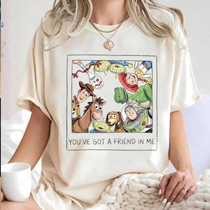 Op de afbeelding: Crèmekleurig T-shirt met een ingelijste illustratie van Toy Story-personages, waaronder Woody, Buzz Lightyear en Rex. De tekst "YOU'VE GOT A FRIEND IN ME" staat eronder. Het shirt is gemaakt van zacht materiaal.
