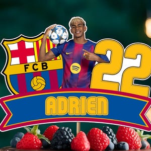 May include: A cake topper featuring a soccer player in a Barcelona jersey, the number 22, and the name "ADRIEN". The cake is decorated with chocolate drips, fresh berries, and the "PREEM GIFTS" logo.