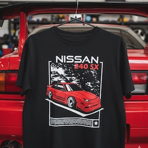 Könnte beinhalten: Schwarzes T-Shirt mit einem roten Nissan 240SX-Autodesign. Das Design enthält den Text "NISSAN 240 SX" in Weiß sowie eine stilisierte Illustration des Autos. Das T-Shirt hängt vor einem roten Auto.