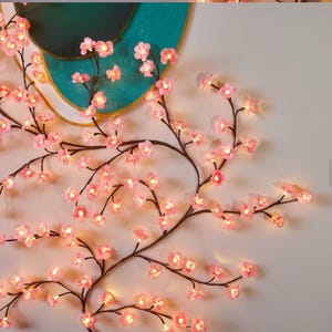 May include: A decorative wall hanging featuring illuminated pink cherry blossom branches. The branches are dark brown and the blossoms are lit with small, warm-toned lights. The background includes a teal and gold circular design.