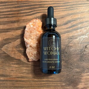 May include: A dark glass bottle with a black dropper, labelled "Witchy Woman Luscious Locks Hair Growth Serum, 2 oz." is next to a cluster of orange crystals. The bottle and crystals are on a wooden surface.