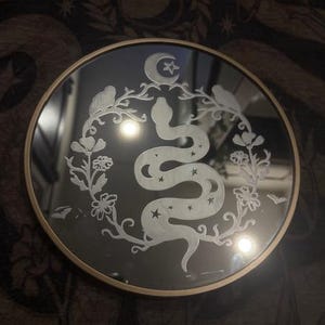 Może przedstawiać: Okrągłe lustro ze złotą ramą. Lustro ma biały, wytrawiony wzór węża, gwiazd, półksiężyca i elementów kwiatowych. Wzór jest wyśrodkowany na powierzchni lustra.