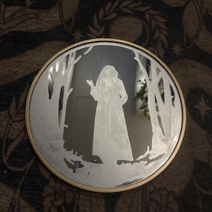 Op de afbeelding: Ronde spiegel met een goudkleurige lijst. Het reflecterende oppervlak toont een silhouet van een persoon in een lange jurk in een boslandschap. Het ontwerp is in de spiegel geëtst.