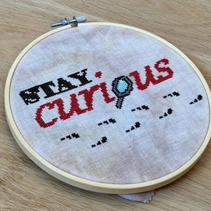 Puede incluir: Un bastidor de bordado circular de madera que contiene tela de color claro con el texto STAY CURIOUS bordado en punto de cruz. La palabra STAY es negra, CURIOUS es roja, con una pequeña lupa azul y gris que reemplaza la letra I.